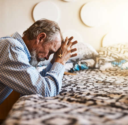 Some time alone to say thanks for everything. Shot of a peaceful senior man kneeling next to his bed and praying with his eyes closed.の写真素材