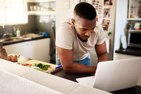 Lets check out some new recipes. Cropped shot of a handsome young man using a digital tablet while making breakfast in his kitchen at home.の写真素材