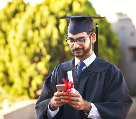 Posting his graduation speech online. Shot of a young man using a mobile phone on graduation day.の写真素材