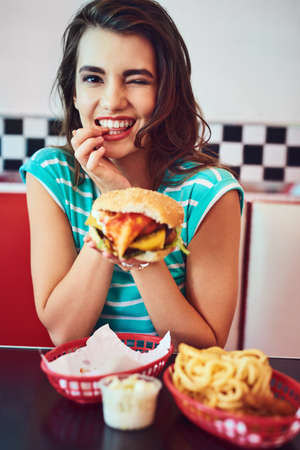 I eat very well. Cropped portrait of an attractive young woman enjoying a burger in a retro diner.の写真素材