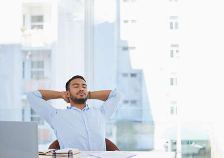 I deserve a break now. Shot of a young businessman sitting with his hands behind his head at his desk in an office.の写真素材
