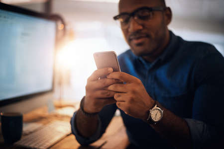 Checking in with a few connections. Shot of a young businessman using a cellphone in an office at night.の写真素材