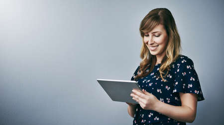 Reading the latest blog post. Studio shot of an attractive young woman using her digital tablet against a grey background.の写真素材