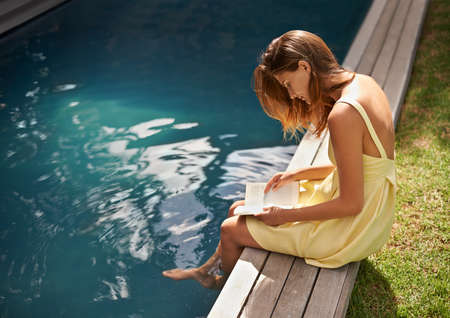 The greatest escapes. An attractive young woman reading her book while resting her feet in the pool.
