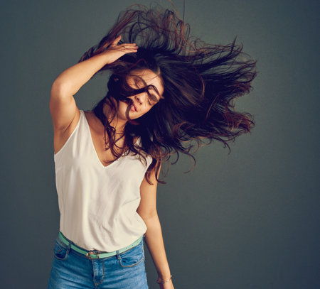Her hair has a mind of its own. Studio shot of a carefree young woman holding her hair while it gets blown by wind as she stands against a dark background.の写真素材