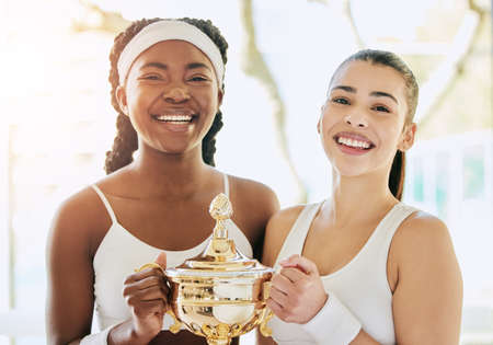 Doubles champions. Cropped portrait of two attractive young female tennis players holding a trophy while standing in the clubhouse during their award ceremony.の写真素材