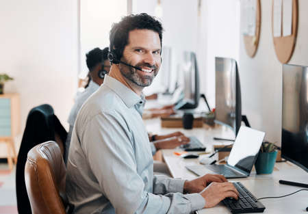 Work as hard as your team does. Shot of a mature agent sitting in the office and using his computer while his colleagues work behind him.の写真素材