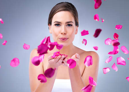 Blossom into a new you. An attractive young woman blowing rose petals against a grey background.の写真素材