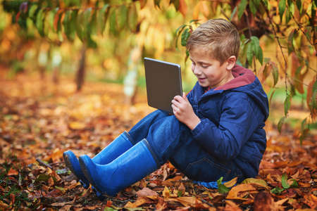 Modern day kid. Full length shot of an adorable little boy using a tablet while sitting outdoors during autumn.の写真素材