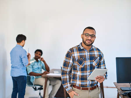 Smart technology makes management so much simpler. Portrait of a young entrepreneur using a digital tablet at work with his team in the background.の写真素材