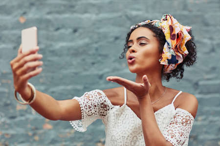 The classic blowing a kiss selfie is a must. Cropped shot of an attractive young woman taking a selfie against a brick wall outside.の写真素材