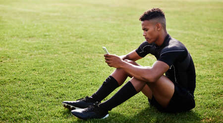 The wifi is as hot as his skills. Shot of a young man sitting on the field and using a smartphone at a rugby match.の写真素材