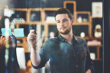 Confident in their plans. Shot of two young businessmen brainstorming on a glass wall in an office.の写真素材
