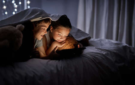 They stay entertained with technology. Shot of two adorable little siblings using a digital tablet together in bed at night.の写真素材