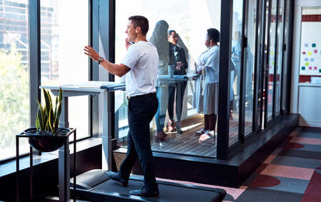 Theres no need to be deskbound as you head to success. Shot of a young businessman talking on a cellphone and going through paperwork while walking on a treadmill in an office.の写真素材