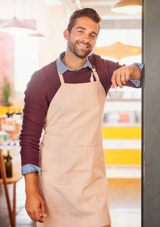 Being my own boss is the best feeling. Portrait of a young business owner standing in the doorway of his coffee shop with an apron on.の写真素材