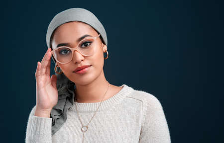 Keeping up with trends. Cropped shot of an attractive young woman standing against a black background alone while wearing spectacles and a headscarf.の写真素材