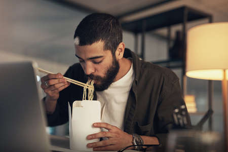 Business gets better with every bite. Shot of a young businessman having takeout and using a laptop during a late night at work.の写真素材