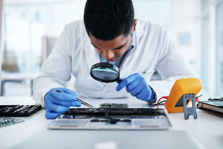 Leave no chip unturned. Shot of a young man using tweezers and a magnifying glass while repairing computer hardware in a laboratory.の写真素材