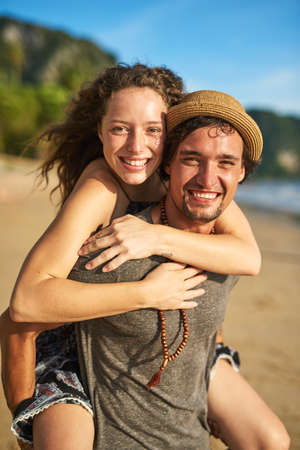 Living our love in the sunshine. Portrait of a happy young man giving his girlfriend a piggyback ride on the beach.の写真素材