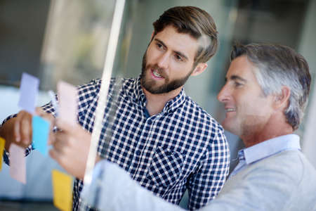 The dynamic duo. Shot of coworkers arranging sticky notes on a glass wall during a brainstorming...