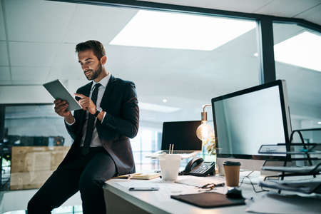 Its only me in the office tonight. Shot of a confident young businessman browsing on his digital tablet while standing inside of the office.の写真素材