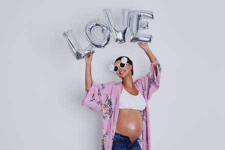 So much love for someone I havent even met yet. Studio shot of a beautiful young pregnant woman holding up a word balloon that reads love against a gray background.の写真素材
