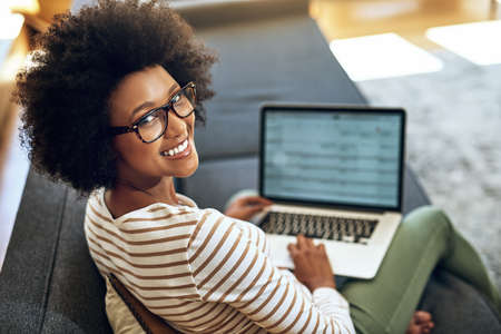 No better way to work than to work from home. Portrait of a cheerful young woman working on her laptop while being seated comfortably on a couch at home.の写真素材