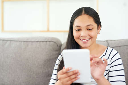 Dipping into the digital world. Shot of a happy young girl relaxing on the sofa and using a digital...