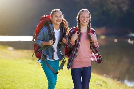 On a girls adventure. Shot of two young girls wearing backpacks walking together beside a lake.の写真素材
