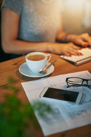 Everything she needs for a productive night at the office. Cropped shot of a woman working on a computer with coffee, a phone and paperwork in the foreground.の写真素材