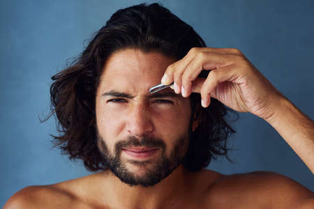 Giving his brows some defined shape. Studio portrait of a handsome young man plucking his eyebrows against a blue background.の写真素材