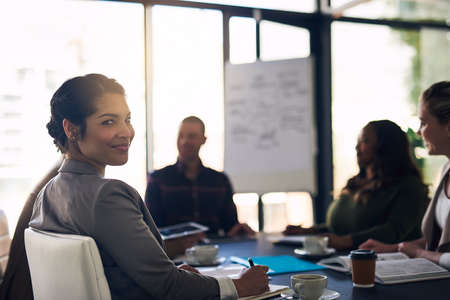 Feeling confident at the meeting. Portrait of a confident business woman sitting in a boardroom meeting with work colleagues while looking into the camera.の写真素材