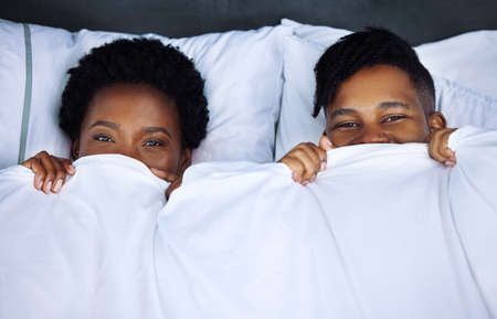 Be back later, going back to bed. Shot of a young couple hiding under the covers in bed at home.の写真素材