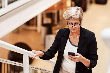 Tending to texts while on the move. Shot of a mature businesswoman using a cellphone while walking up a staircase in an office.の写真素材