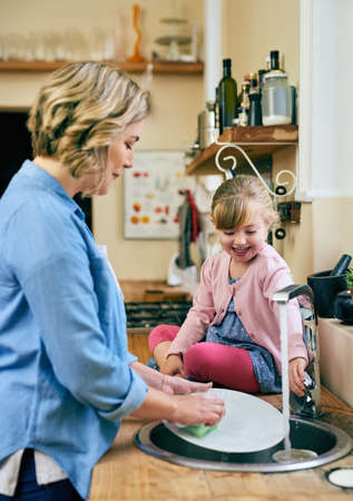 Teaching her daughter the domestic basics. Shot of a cheerful young mother and her young little daughter washing dishes together at home.の写真素材
