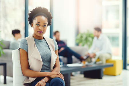She has an attitude for excellence. Portrait of a confident young creative sitting in an office with colleagues in the background.の写真素材