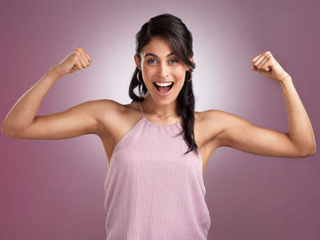 You are beautiful, you are strong, you are worth it. Shot of a beautiful young woman looking cheerful and flexing her arms while standing against a pink background.の写真素材