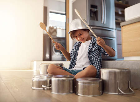 Hes a born percussionist. Shot of a happy little boy playing drums with pots on the kitchen floor while wearing a bowl on his head.の写真素材