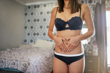 Love your body. Shot of an attractive young woman in underwear making a heart shape on her stomich...