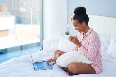 Going through my blog before my day starts. Shot of an attractive young woman drinking coffee while using her laptop in bed at home.の写真素材