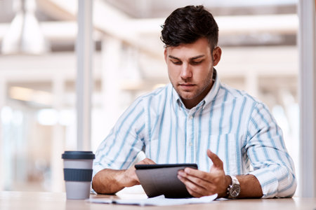 Doing his business wirelessly. Cropped shot of a handsome young male designer using his tablet while sitting in the office.の写真素材