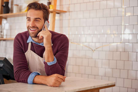 Checking in with his suppliers. Shot of a happy business owner using his smartphone while leaning on the counter in his coffee shop.の写真素材
