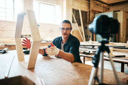 My DIY videos are always trending online. Shot of young carpenter recording a tutorial of him making a wooden masterpiece on a video camera inside his workshop.の写真素材