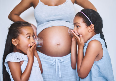 Theres movement in there. Shot of two cheerful little girls standing next their mother while each putting a glass on her pregnant belly to listen for movement at home.の写真素材
