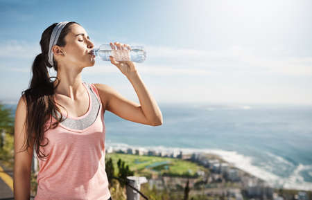 Filling up after a good run. Cropped shot of a young woman drinking a bottle of water while out...