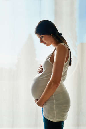 Motherhood is her miracle. Shot of a pregnant woman cradling her belly while standing in front of a window at home.の写真素材
