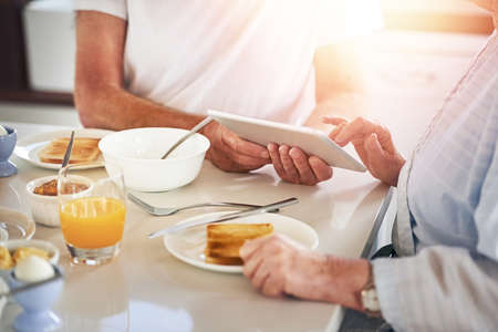 Browsing at breakfast. Closeup shot of a senior couple using a digital tablet while having breakfast at home.の写真素材