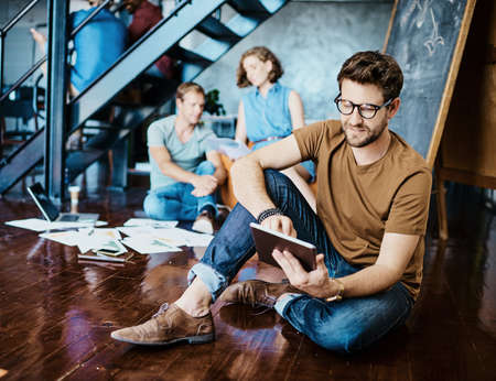 Using smart tools to gain a competitive edge. Shot of a young designer working on a digital tablet with his colleagues in the background.の写真素材