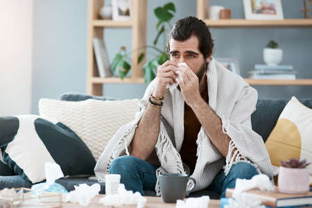 This is not how I planned on spending my weekend. Shot of a sickly young man blowing his nose with a tissue in his living room.の写真素材